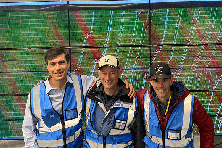 Three researchers wearing blue safety vests pose in front of a large screen that shows a tangle of red, green, and white lines.