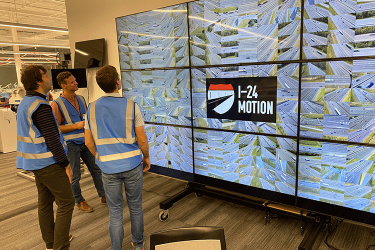 Three researchers wearing blue safety vests stand in front of a grid of nine screens that is approximately nine feet tall. The center screen reads “I-24 motion.”