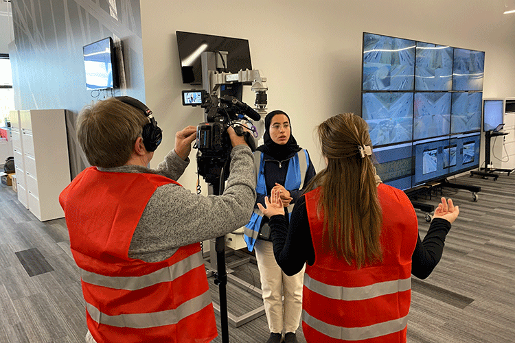 A researcher wearing a blue vest over her hijab speaks to two journalists wearing red vests.