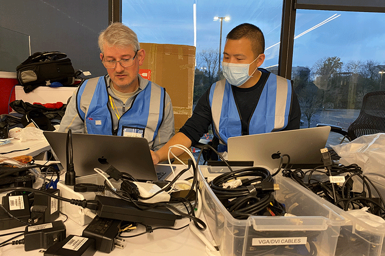 Two researchers wearing blue safety vests talk to each other while working on laptops.