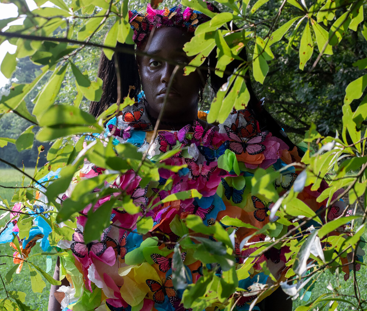 a person wearing a costume made of fake butterflies stands outside behind the leaves of a tree