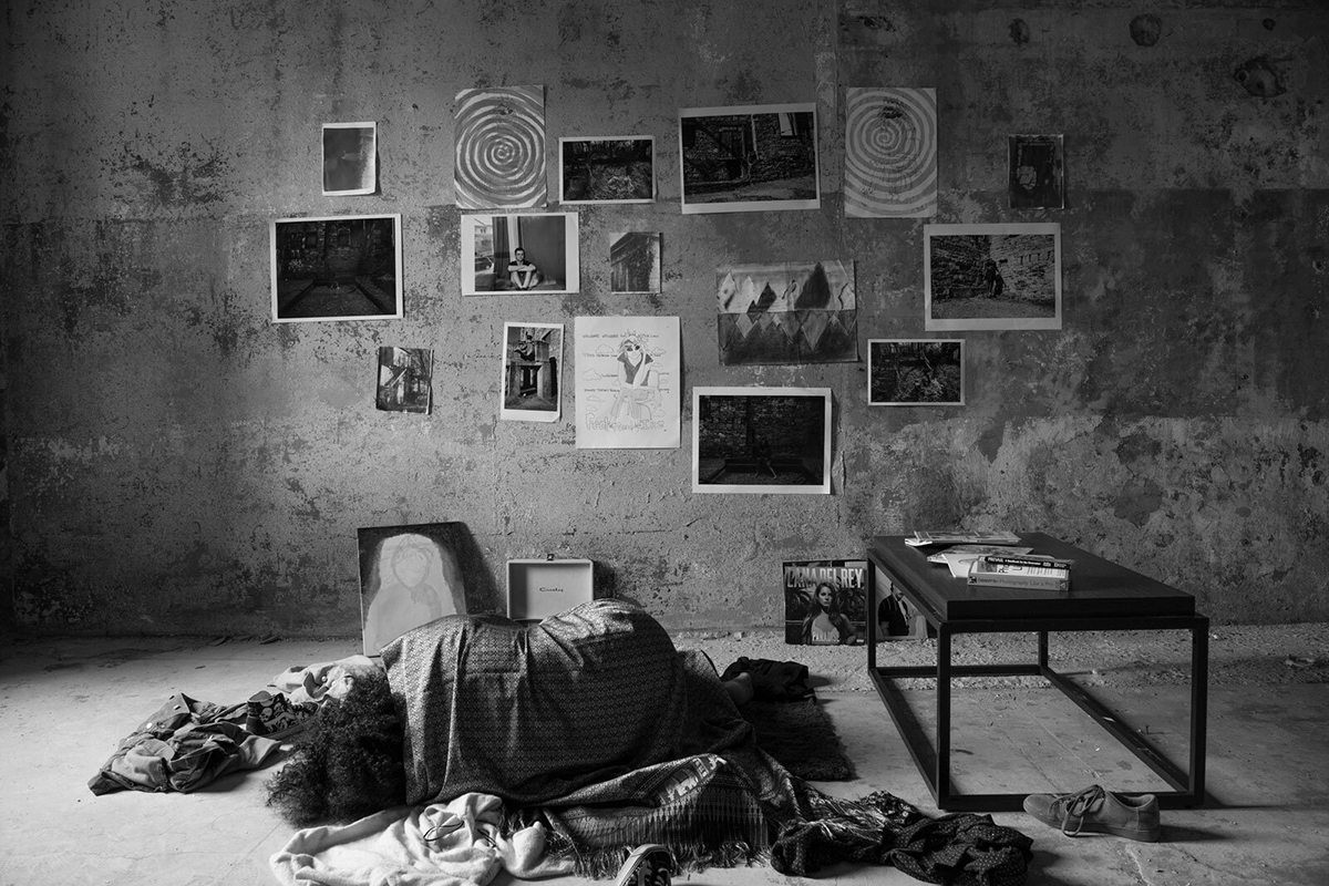 a person lies on a concrete floor with a scarf covering her body and photos taped to a concrete wall