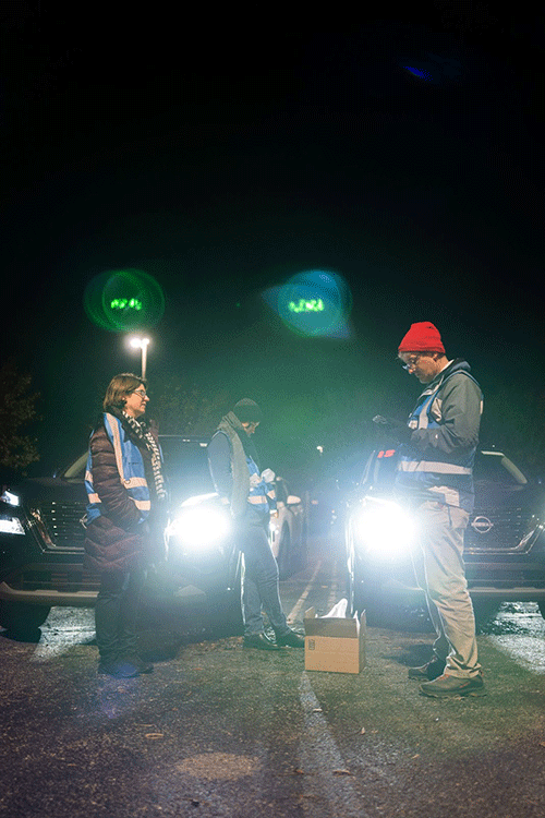 Three researchers wearing blue safety vests stand in a parking lot at night.