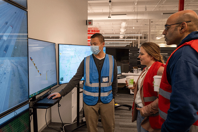 A photo shows a researcher in a blue vest showing an image on a computer screen to two individuals in red vests.