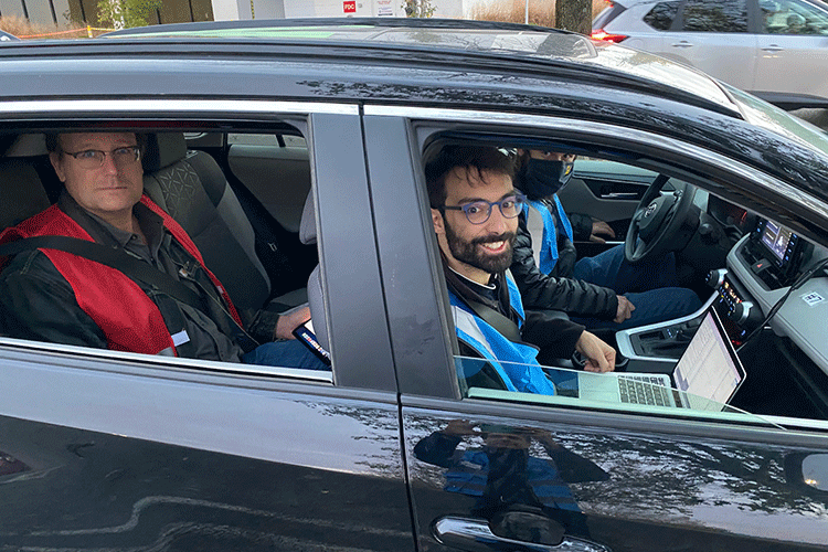 A photo shows two people in blue vests sitting in the front of a car and one person in a red vest in the back of a car.