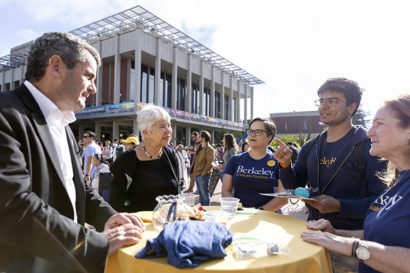 Lisa García Bedolla talks with people at an outside table during a sunny day, there's a crowd of people behind her on Sproul Plaza