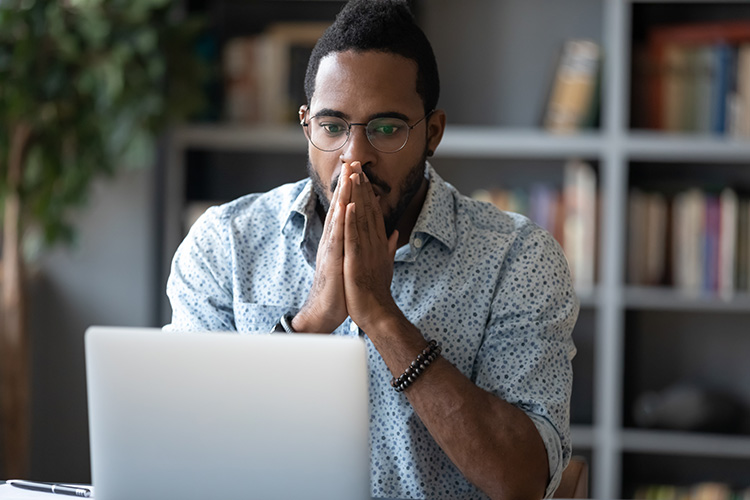 A man sitting in front of a computer screen holds his hands together as if viewing something very traumatic. looking stressed at a laptop