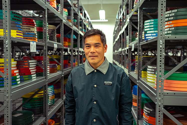 Jon Shibata stands at BAMPFA's Richmond Field Station film vault with rows of colorful film cans on either side of him. He is looking straight at the camera.