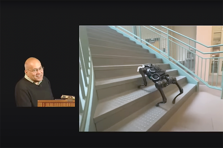 a person giving a lecture watches a video of a robot walking upstairs