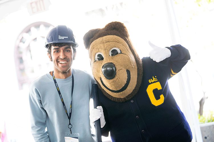 An undergraduate student wearing a hard hat and a lanyard poses next to a large bear mascot