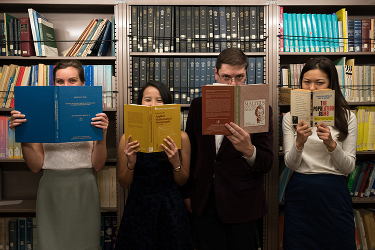 four people holding books in front a bookshelf, looking at the camera