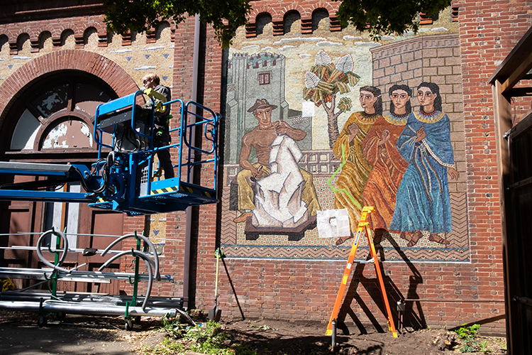 A man on a cherry picker starts work repairing the mosaic murals on the Old Art Gallery building on campus.