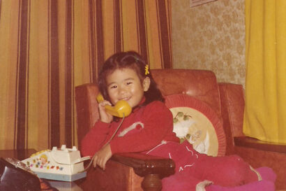 Sunny Lee as a young child sits in a chair smiling while holding a yellow phone