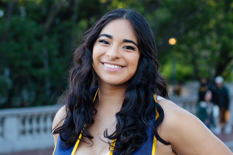 informal portrait of Andrea Sandoval, wearing her graduation stole, smiling, against a campus backdrop of low balustrades and trees
