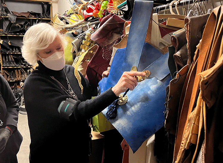 Inside a closet storage room that is filled with different pieces of costuming, a woman wearing a face mask holds a bright blue piece of costume chest armor and points to a detail in the material.