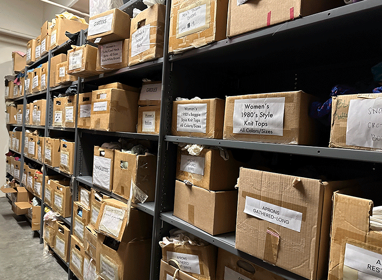 A photo shows racks of cardboard boxes that are stacked floor to ceiling along the wall of a large storage room. Each box is labeled with a piece of paper that has been taped on. One label reads “Women’s 1980’s Style Knit Tops All Colors/Sizes.” Another reads, “Aprons, Gathered – Long.”