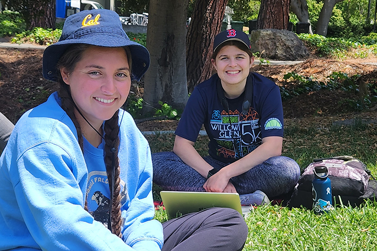 UC Berkeley students Violet Henneberger and Annie Zuromski, who also are Fledge Watch volunteers, sit on the grass near the Campanile and smile at the camera. Violet is wearing a Cal hat and a Cal Falcons sweatshirt and Violet has her laptop open and a water bottle and backpack next to her.