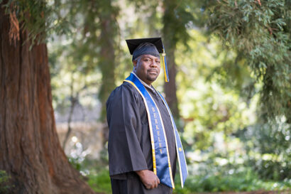 man in black graduation gown with cap, looking at camera while standing in grove of trees