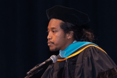 Justin Jones, in commencement attire, stands against a black background and delivers the keynote address at the Goldman School of Public Policy commencement ceremony