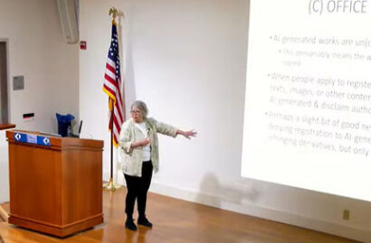 Pam Samuelson stands on stage talking while pointing to a slide that is projected onto the wall