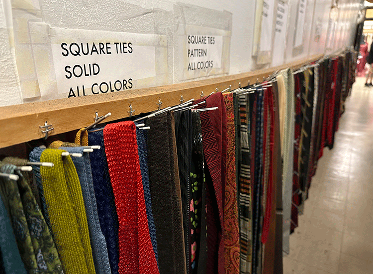 A photo shows a rack of hanging ties of all colors that extend along the wall of a storage room. A paper sign taped above the ties reads “Square Ties, Solid, All colors.”