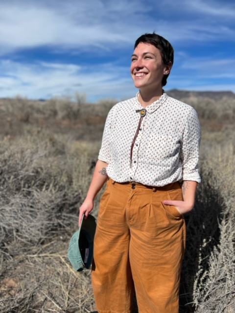 a person with short hair wearing a light-weight collared shirt and orange pants stands in a field and smiles