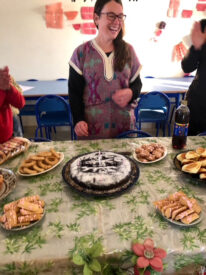 a person wearing glasses and colorful clothes and smiling stands in front of a table of cakes and other deserts