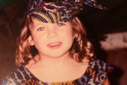 a young kid with long hair wears a hat and dress, both made of colorful African fabric