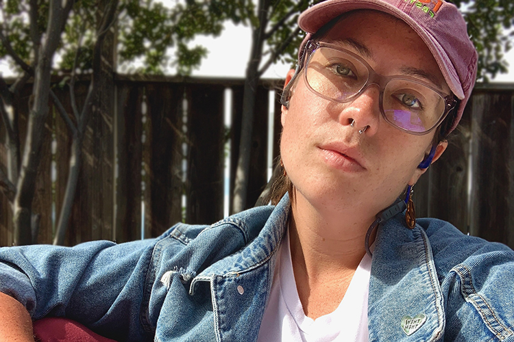 a person wearing a hat and jean jacket sits outside resting their arm on their knee and looks straight into the camera with a serious look on their face