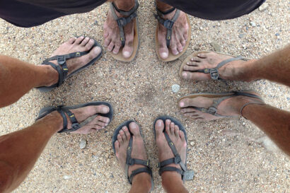 a close-up photo of four people's feet wearing sandals standing on a dirt and rocky clear