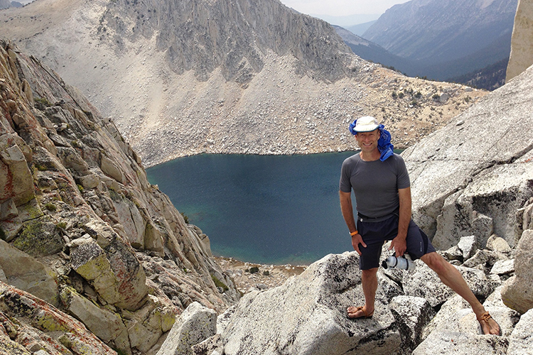 a man in a grey shirt and white hat wearing sandals, standing on rocks in the mountains above a lake