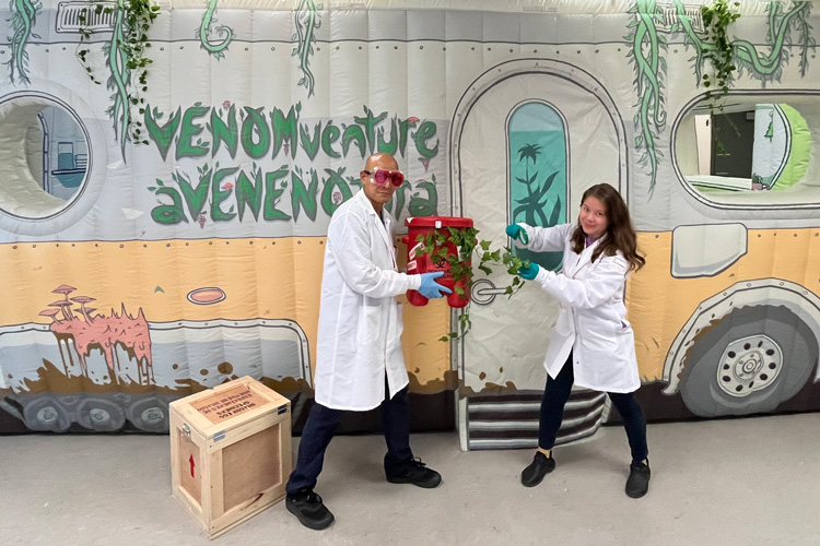 a man and woman in white lab coats carry a pail filled with a vine in front of an inflatable designed to look like a mobile lab. VENOMventure is written on the side.