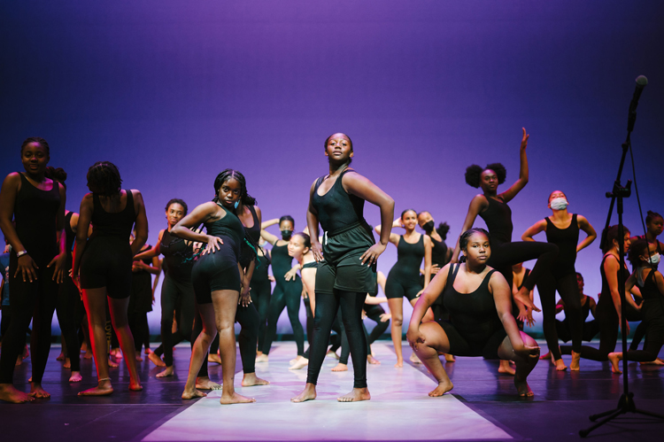 a few dozen young dancers dressed in black leotards strike poses on a fashion show runway