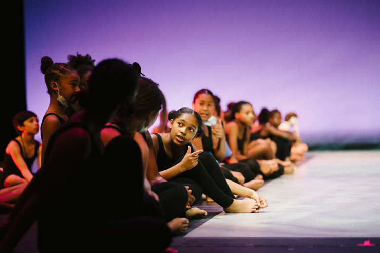 Young dancers in black leotards sit in a row on the floor, talking to each other