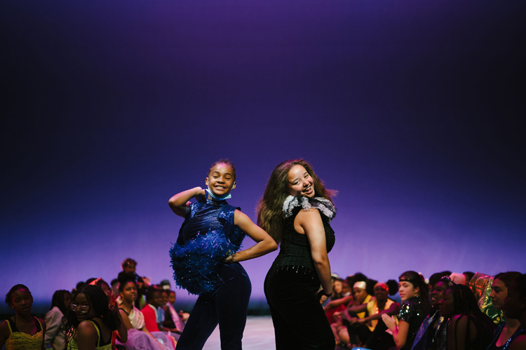 Two young dancers wearing costumes pose at the end of a runway as dancers lining both sides of the runway watch