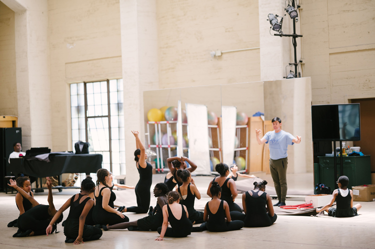 About a dozen young dancers sit on the floor of a gym with a lot of windows as a teacher stands and explains something to them