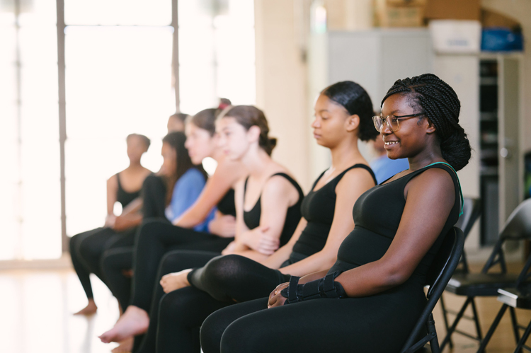 Several young dancers wearing black leotards sit on a chairs in a row watching something out of the frame