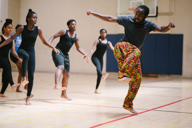 A dancing teacher wearing coloring pants teaches a dance to several young dancers in a gym