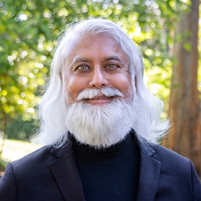 headshot of a person with light eyes, white shoulder-length hair and a white beard standing outdoors and smiling