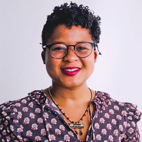 headshot of a person smiling with short, curly dark hair wearing glasses, dark pink lipstick and a patterned shirt