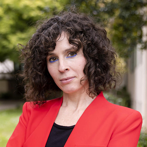 headshot a person with dark brown curly hair and a bright red-orange blazer smiling slightly