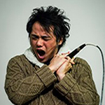 a person with short spiky hair and wearing a chunky green sweater sings to the side and holds a mic in their hands