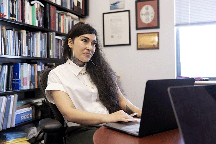 A photo of a person sitting at a desk in an office and looking at a laptop. She has long dark hair and is wearing a white shirt.