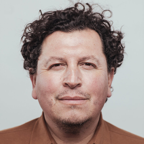 headshot of a person with dark brown, short curly hair smiling slightly