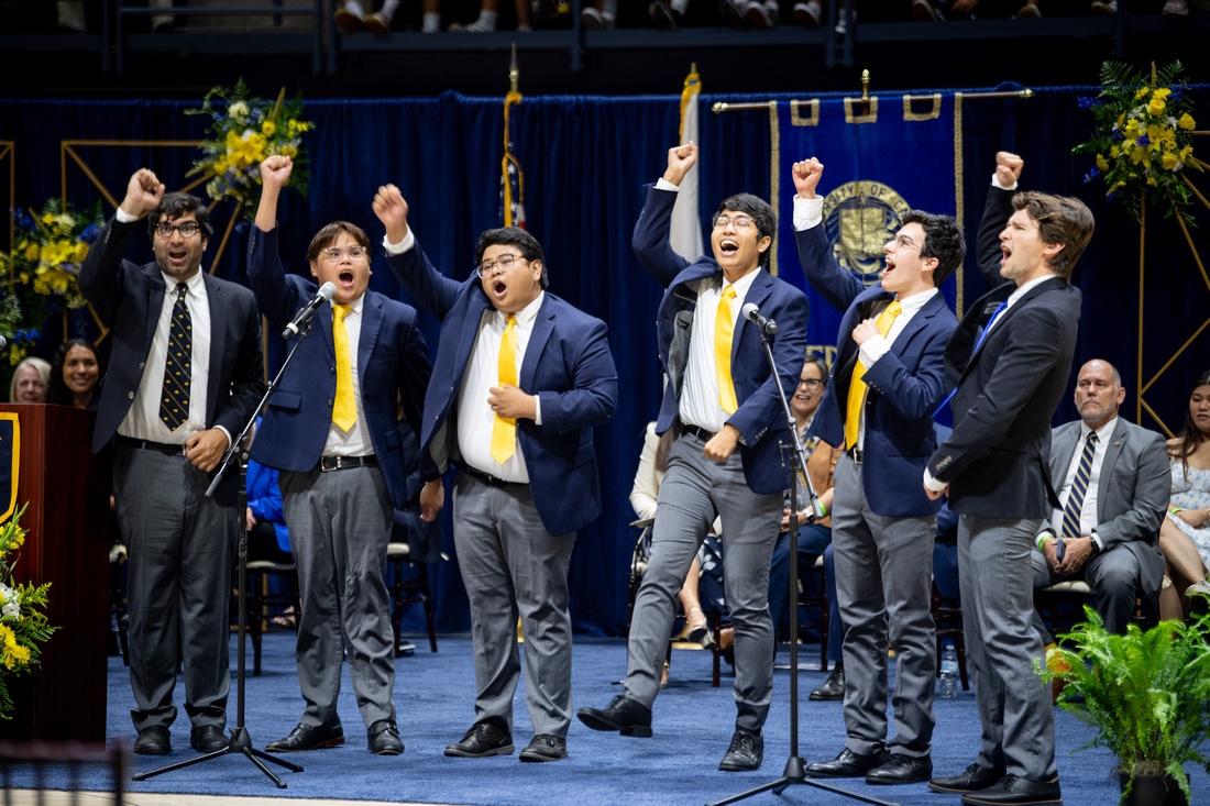 a group of people wearing gray pants, blue jackets and yellow ties raise their arms while singing