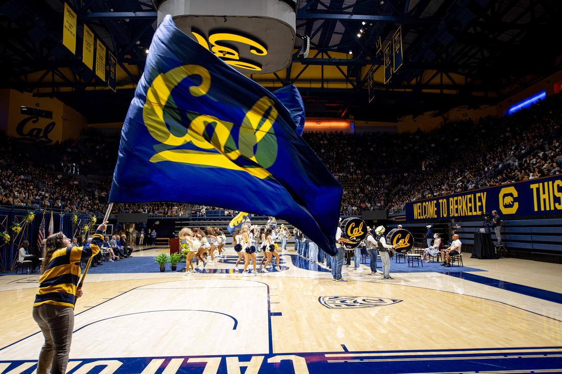 a person waves a blue flag that says Cal while a marching band performs on a gymnasium floor