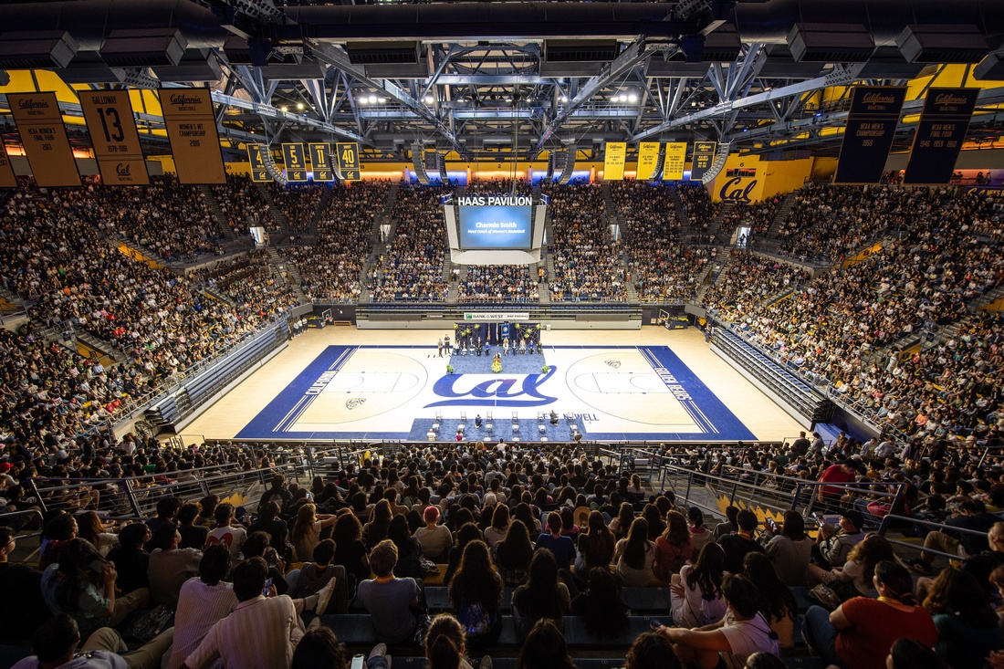 a picture of thousands of students seated in a gymnasium watching people speak on the basketball court