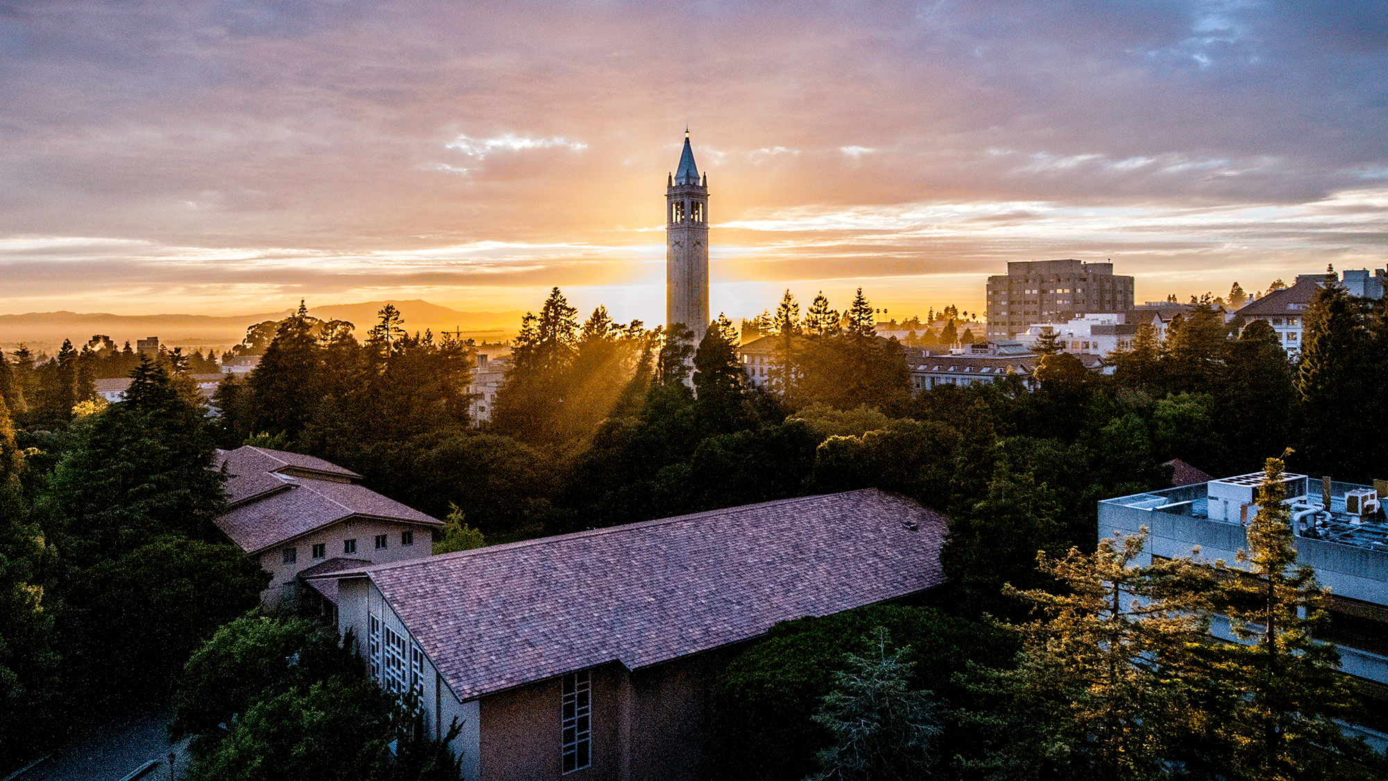 A photo overlooking the rooftops of the Berkeley Campus at dusk. In the center of the photo, the sun is setting behind the campanile.