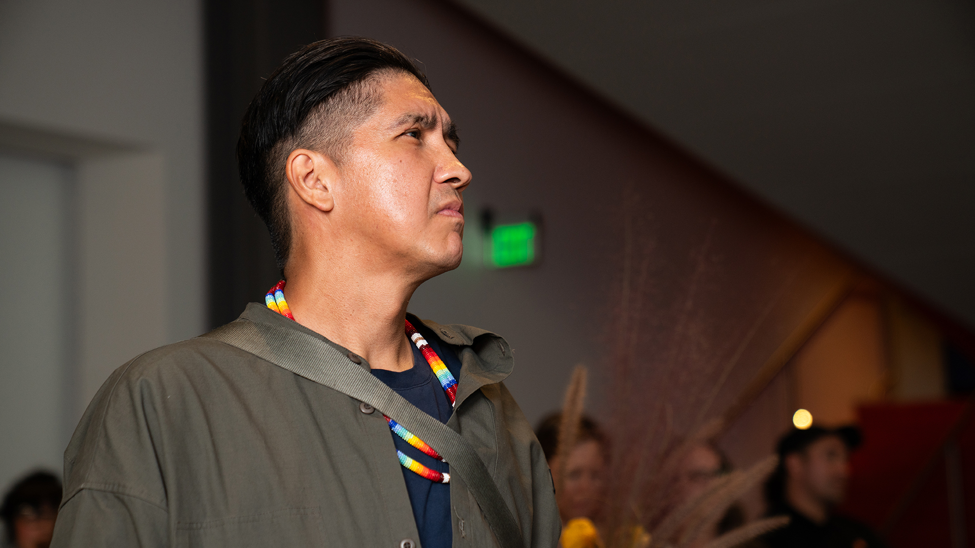 A person with short dark hair shaved on the sides wearing a colorful beaded necklace looks into the distance with a serious look on his face in a room with other people