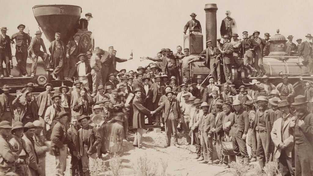 a sepia toned photo of about 100 white men in hats shaking hands and celebrating, some of whom are on top of a train.
